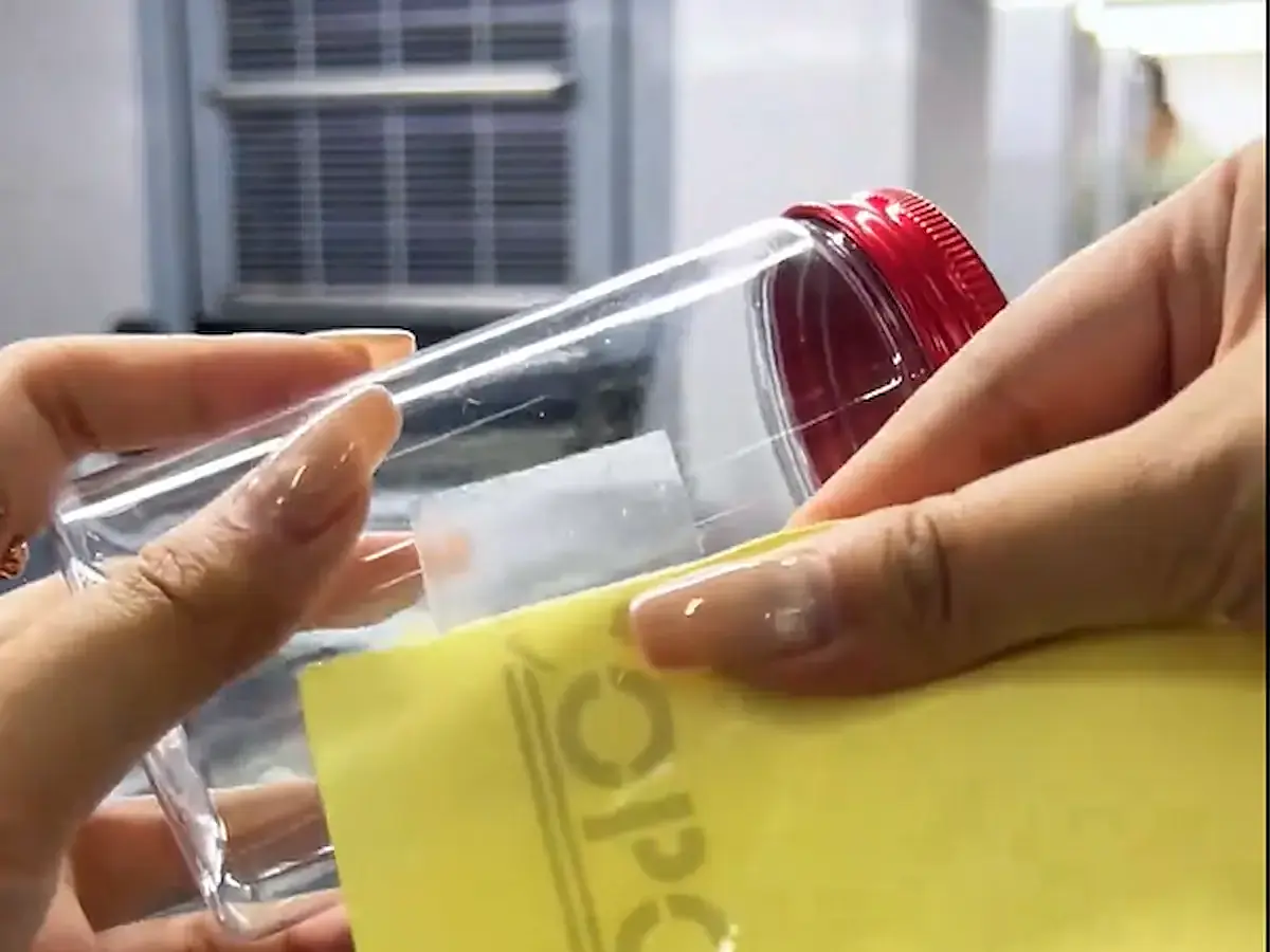 Hands place a clear label on a transparent bottle while holding the yellow backing sheet.