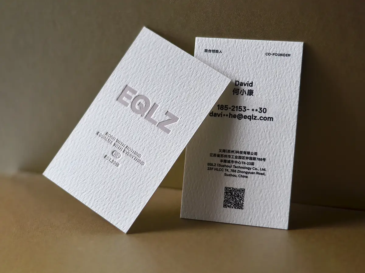 Two white textured cotton paper cards with raised lettering, printed contact details, and a QR code.